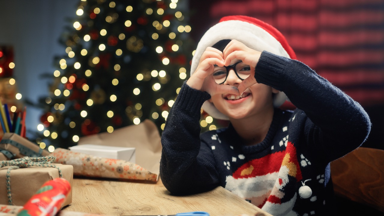 Kleiner Junge vorm Weihnachtsbaum mit Weihanchtsmütze formt mit seinen Händen ein Herz
